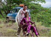 The lost city: a woman in a pink suit tied to a chair with a man wearing white trying to move her and the chair. They are in a jungle and there is a small car behind them.