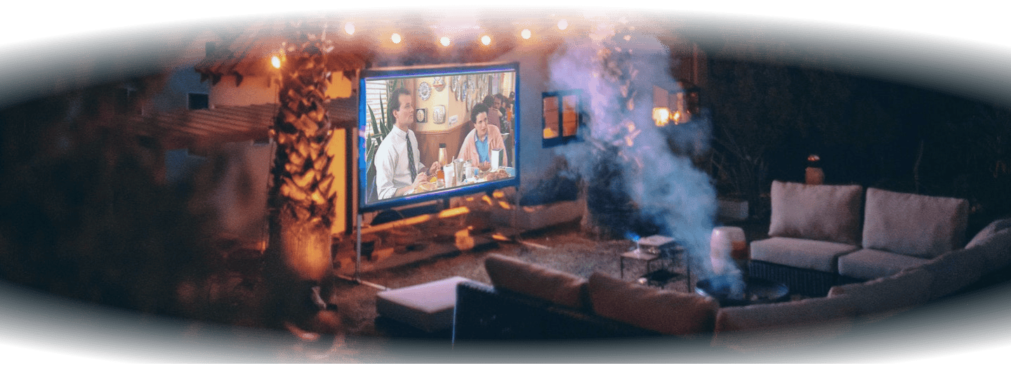 A huge canvas with a movie playing in front of a house and inbetween two palm trees that have a string of lights between them as well, and a huge couch in front of the canvas.